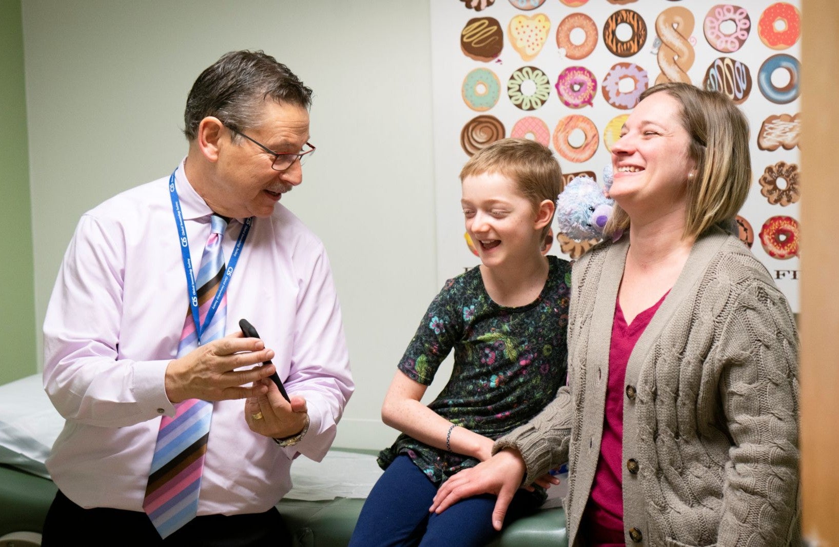 A man holds cell phone up as a child and mother smile. The man is wearing a light pink button up and colorful tie. The child has short hair, a dark colorful shirt, and blue pants. The mother wears a pink shirt and beige cardigan. 