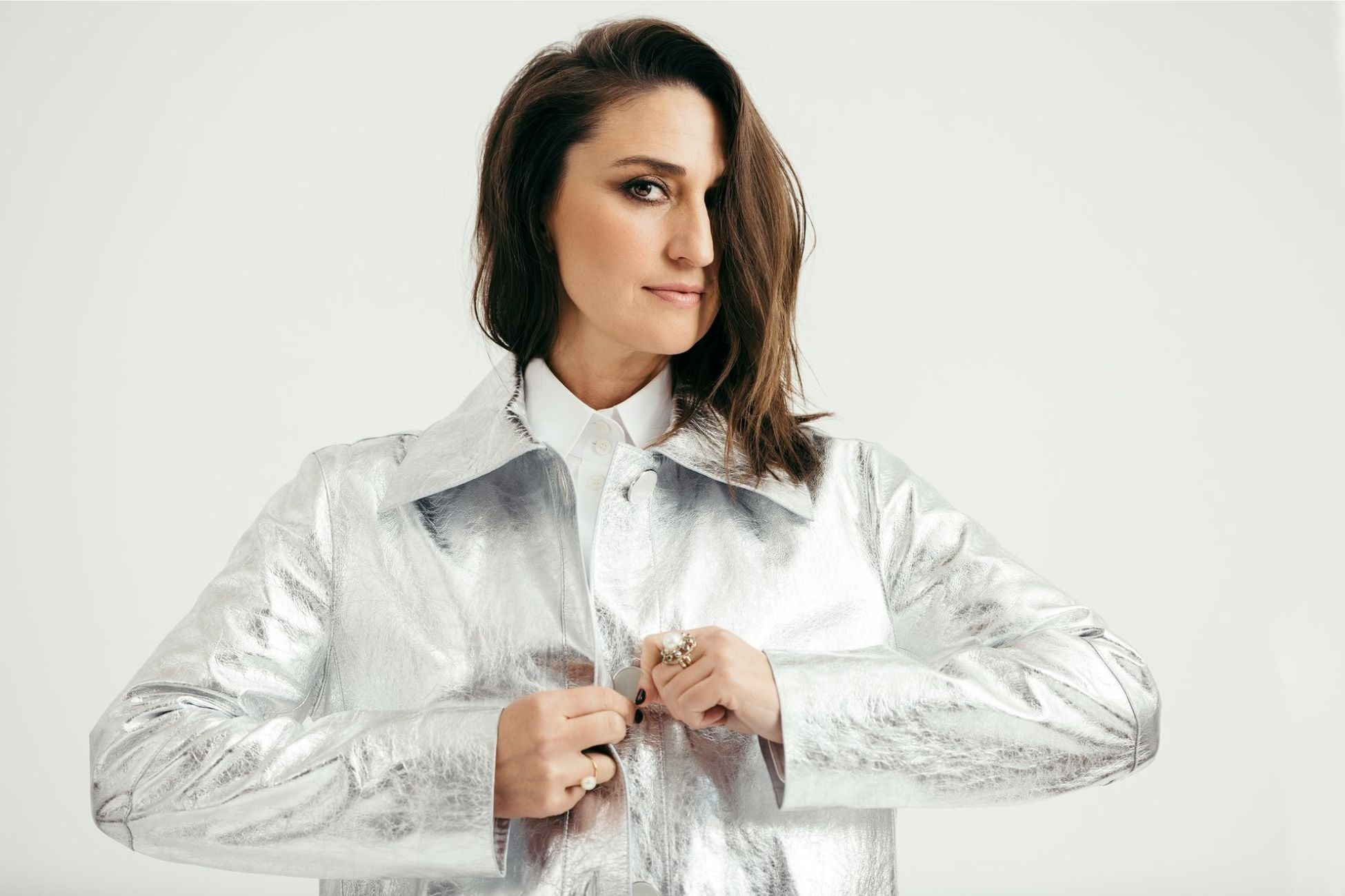 A woman buttoning a shiny jacket over white shirt while smiling confidently at the camera