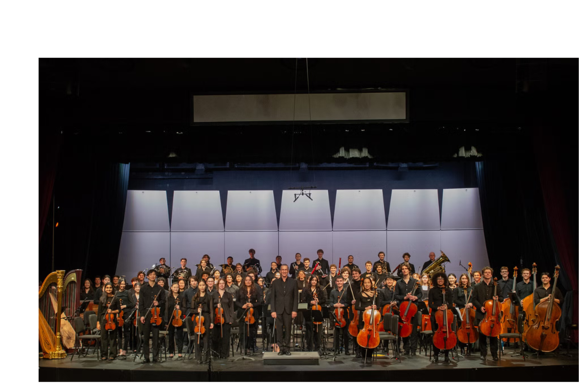 A large university philharmonic orchestra poses onstage in a concert hall after a performance. Musicians dressed in black hold violins, cellos, basses, brass instruments, and a harp, with the conductor standing at the center front beneath bright stage lighting.