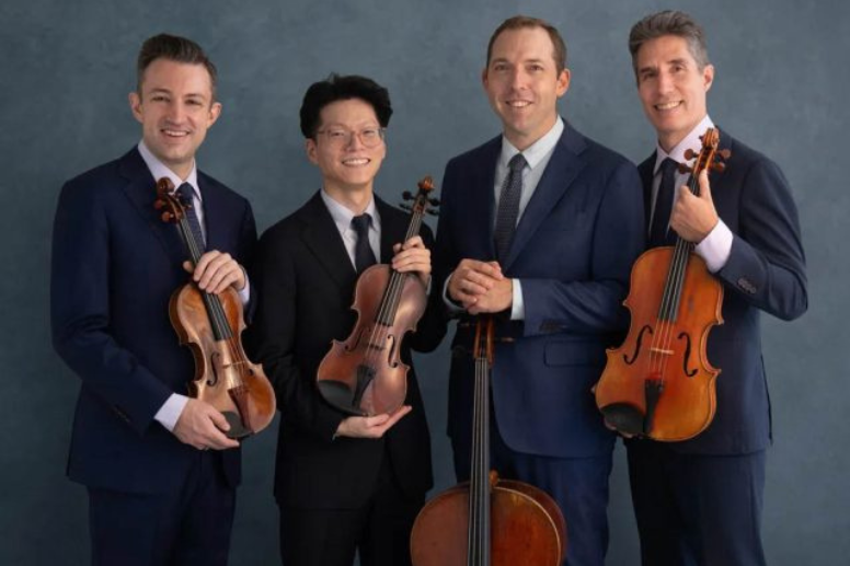 Four musicians in suits pose together holding violins and a cello against a blue-gray background.