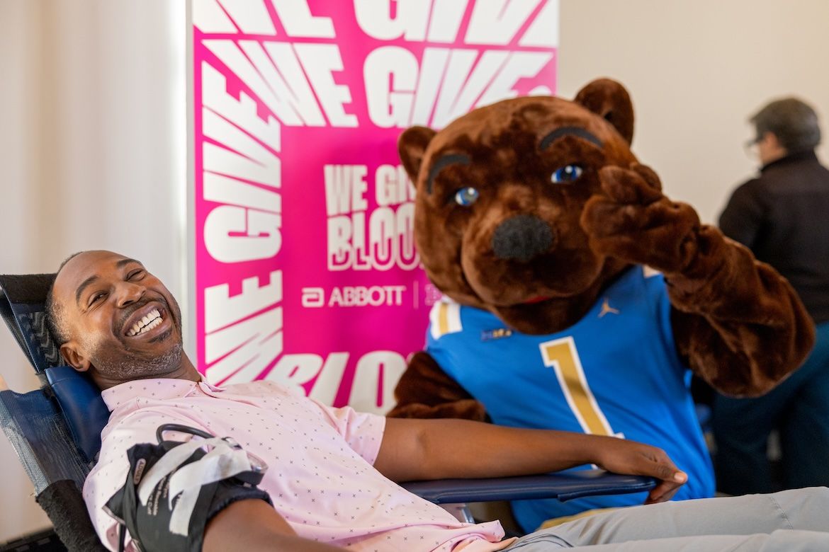 UCLA Athletic Director Martin Jarmond reclines in a blood-donation chair with a bandage on his arm as Joe Bruin, wearing a UCLA jersey, stands beside him and gives a thumbs-up.