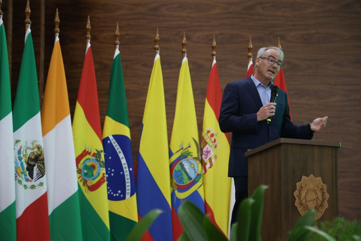 William Boyd stands behind a wooden podium in front of large flags from countries around the world