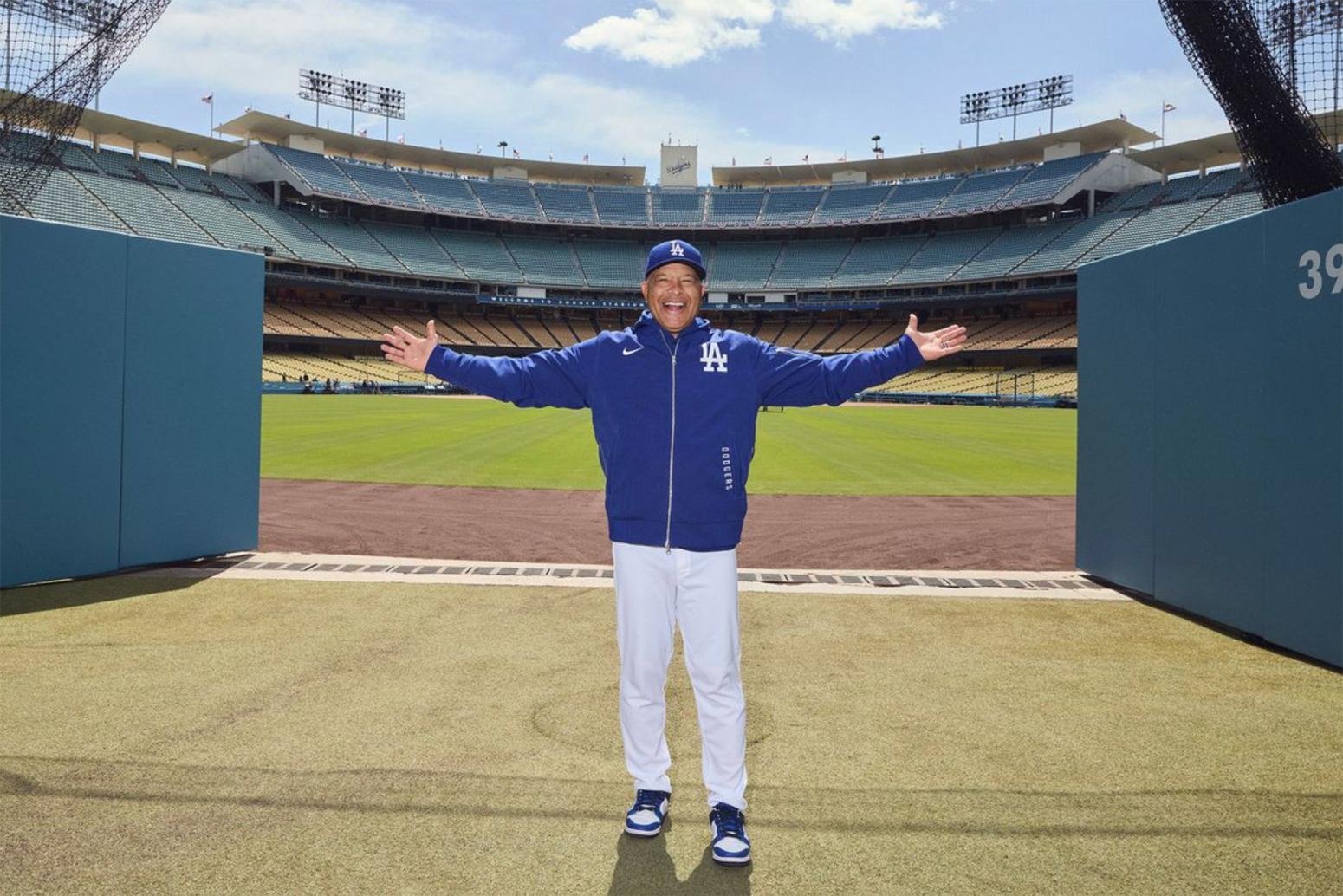 Dave Roberts, in blue LA zip hoodie and cap and white pants, stands arms outstretched with Dodger Stadium behind him